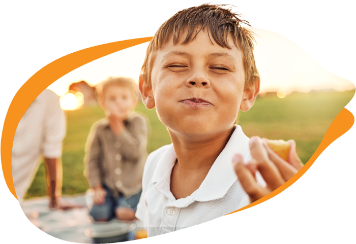 Um menino com os olhos fechados e uma expressão de prazer, mastiga algo que segura com a mão direita. Ele está ao ar livre em um dia ensolarado, com outras pessoas em segundo plano desfocado.